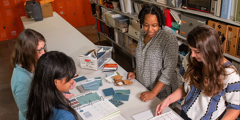 A female design professor stands alongside three female students as they review color and design swatches for a class project. The narrow worktable has color samples – blue, gray, mauve – spread across it.